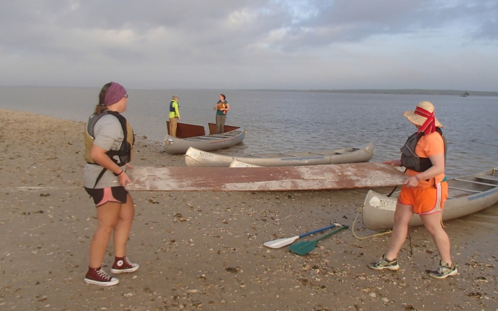 In the foreground, two people carry what appears to be a panel of wood across a beach. Behind them, canoes rest on the shore.